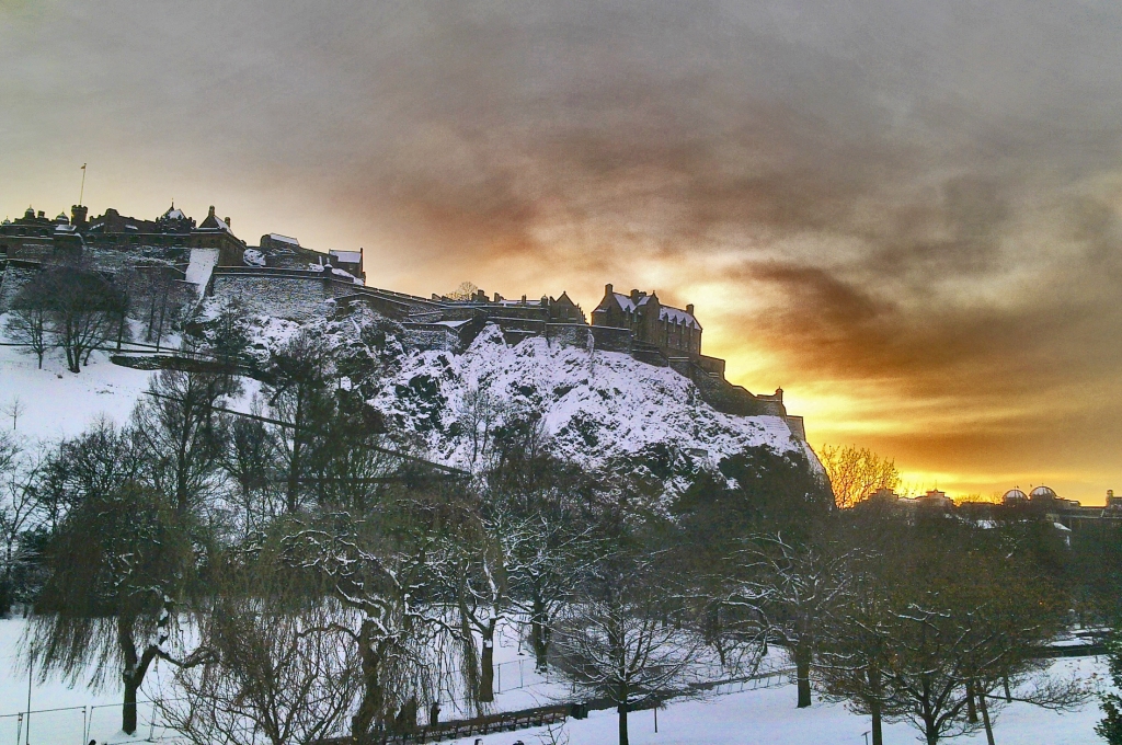 View from Princes Street of Edinburgh Castle covered in snow with a sunset in the background.
