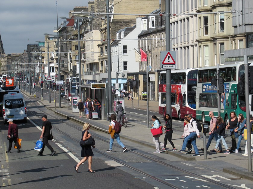 People crossing the road and tram lines at a busy crossing point on Princes Street. Buses & shops are in the background.