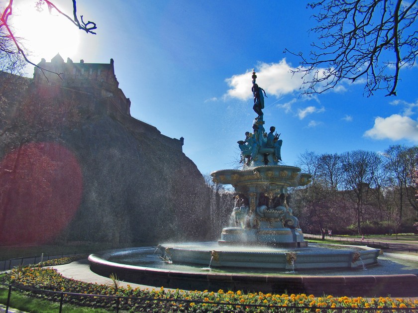 The Ross Fountain on a bright, sunny day. Plants and greenspace surround the fountain. Trees are in the background and a silhouette of Edinburgh Castle sits above.