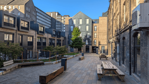 A new development in the city centre on a bright sunny day. Public space shows benches, seating and trees with tall offices in the background.